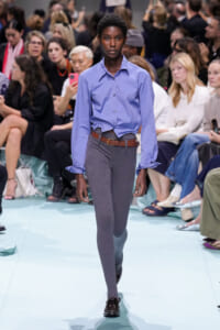 Model walks runway wearing a light blue button-down shirt, gray ribbed tights, brown belt, and black loafers. Audience in background.