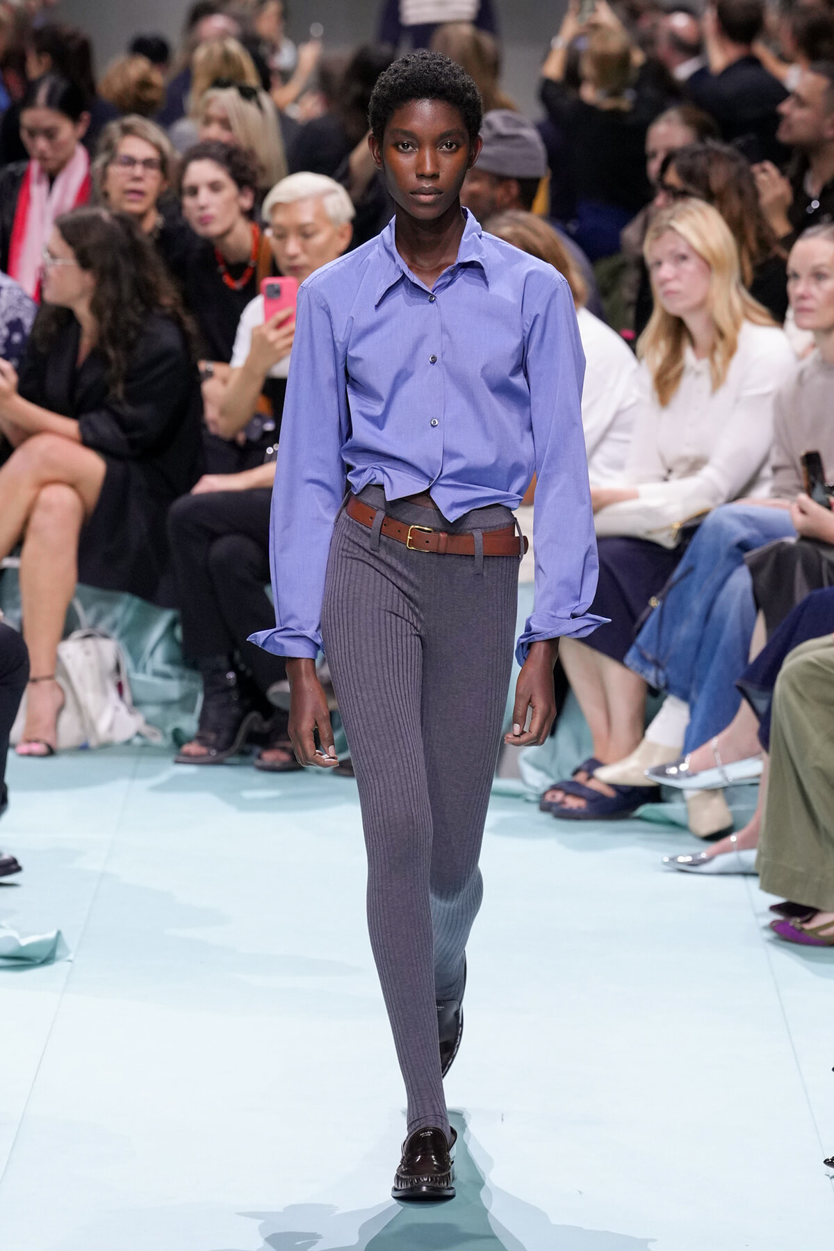 Model walks runway wearing a light blue button-down shirt, gray ribbed tights, brown belt, and black loafers. Audience in background.