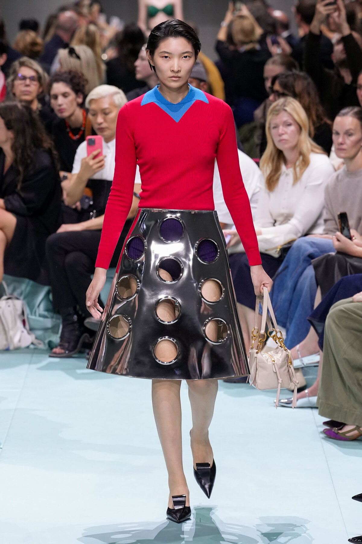Model walks the runway in a red knit sweater with a blue collar and a glossy black circular-hole A-line skirt, carrying a pale handbag.