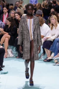 Model walks a fashion runway in a long textured gray coat over a pastel striped bodysuit, audience seated along the catwalk.