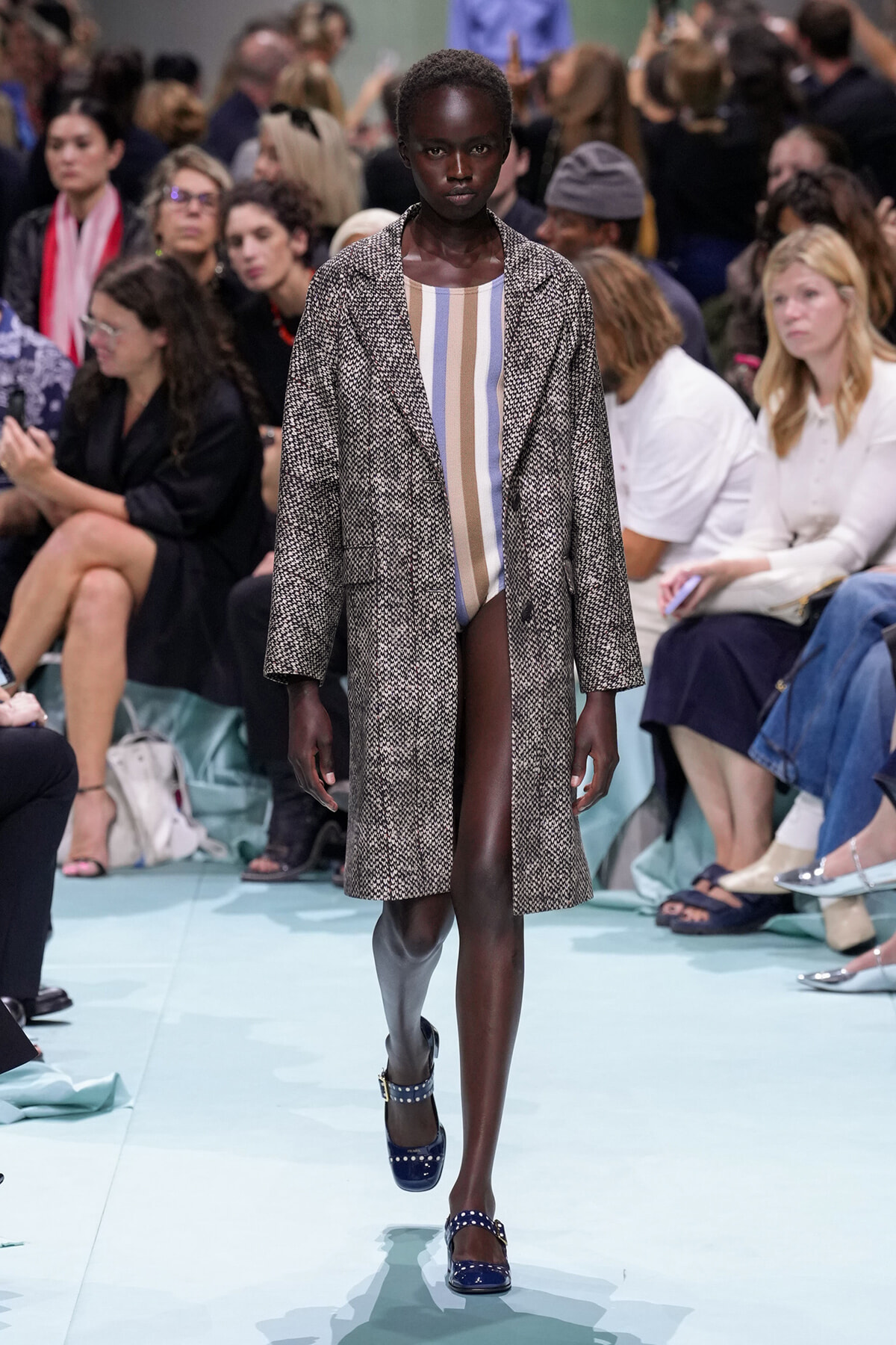 Model walks a fashion runway in a long textured gray coat over a pastel striped bodysuit, audience seated along the catwalk.