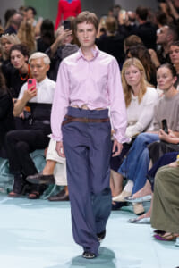 Model walks a fashion show runway in a light pink cropped shirt tied at the waist and navy wide-leg trousers with a brown belt; audience sits along the catwalk.