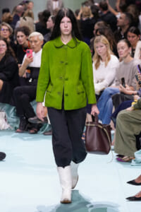 Model walks the runway in a bright lime-green button-front jacket, black pants, white boots, carrying a dark brown tote.