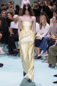 Runway model in a pale yellow strapless satin gown with a gathered skirt and oversized geometric sunglasses.