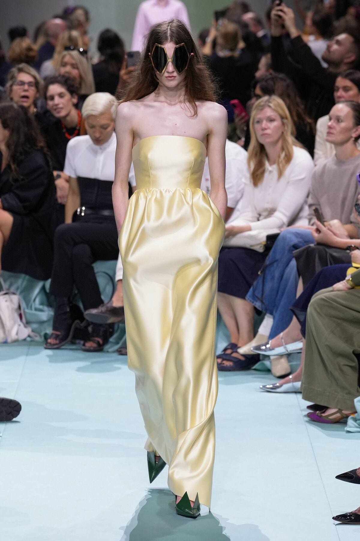 Runway model in a pale yellow strapless satin gown with a gathered skirt and oversized geometric sunglasses.
