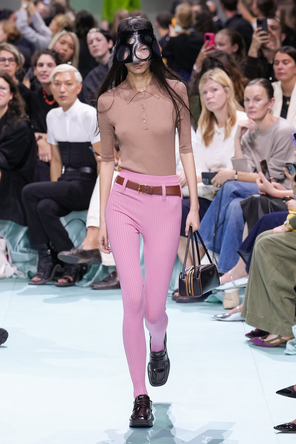 Model walks a fashion show wearing pink ribbed pants, a tan brown top, brown belt, black handbag, and an oversized black mask; an audience watches.