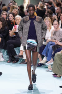 Black female model walks the runway in a textured gray oversized coat over a navy top and metallic silver mini skirt, with black loafers.