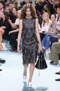Model on a runway wearing a sleeveless dark textured dress covered in metal rings, carrying a black handbag.