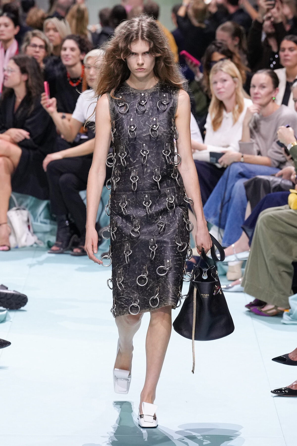Model on a runway wearing a sleeveless dark textured dress covered in metal rings, carrying a black handbag.