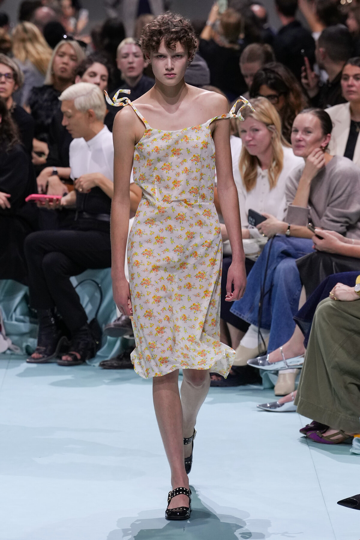 Model walking a runway in a white floral midi dress with tied shoulder straps, audience in the background.