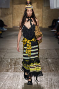 Model walks a runway in a layered outfit: black lace cami, yellow plaid shirt tied at the waist, a striped tiered skirt, knit hat, and tan bag.