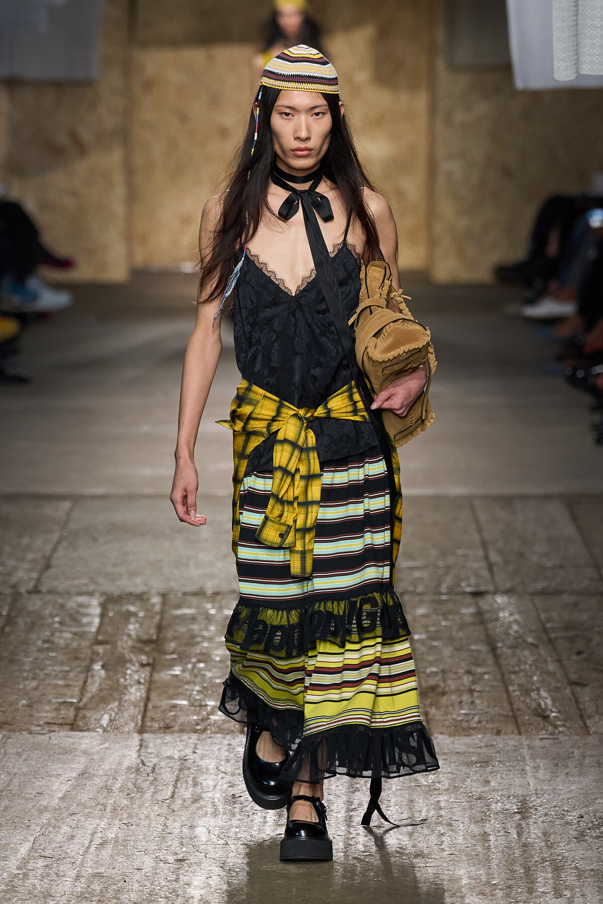 Model walks a runway in a layered outfit: black lace cami, yellow plaid shirt tied at the waist, a striped tiered skirt, knit hat, and tan bag.