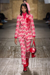 Model struts down a fashion runway in a pink and white graphic suit with red shirt, red boots, and a round red handbag.