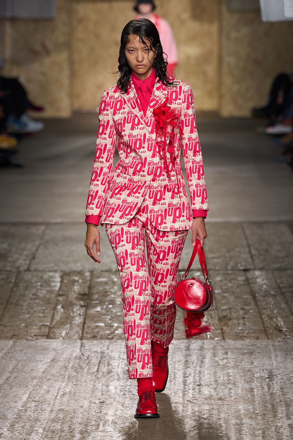 Model struts down a fashion runway in a pink and white graphic suit with red shirt, red boots, and a round red handbag.