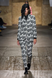 Model on a runway wearing a black-and-white suit with a repeated 'What’s Up!' text pattern and a black textured hat obscuring one eye, walking confidently.