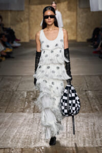 Runway model in a white fringe, polka-dot dress with long black gloves and oversized sunglasses, carrying a black-and-white checkered bag.