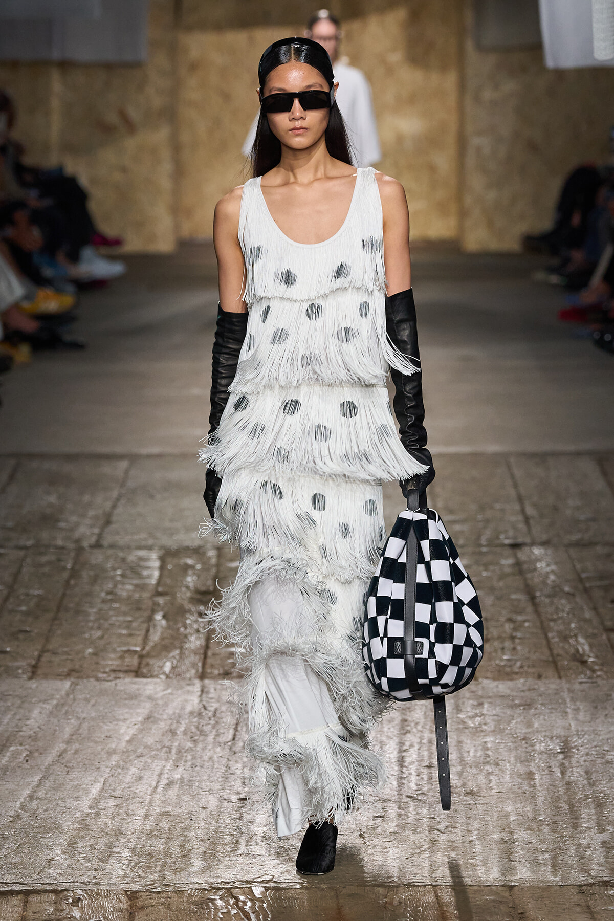 Runway model in a white fringe, polka-dot dress with long black gloves and oversized sunglasses, carrying a black-and-white checkered bag.