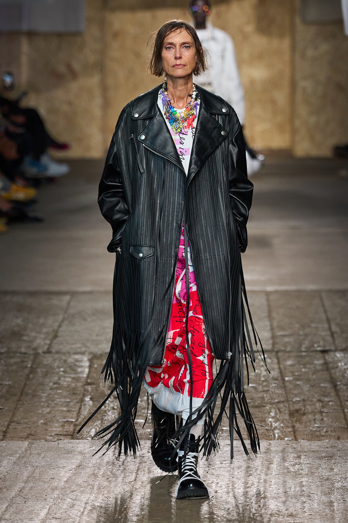 Model walking a runway in a long black fringed leather coat over a bright pink-print dress, accessorized with a colorful layered necklace and black lace-up boots.