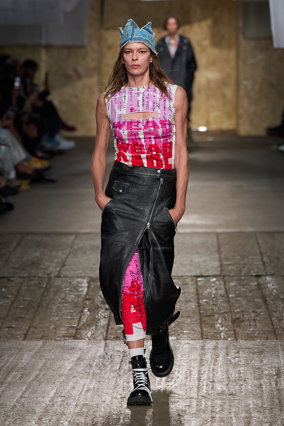 Model on a runway wearing a denim crown, a sleeveless pink/red print top, a black asymmetric leather skirt with a front zipper, pink patterned leggings, white socks, and black lace-up boots.