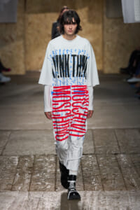 Runway model walks in an oversized white top with bold black lettering and red graphic text, paired with white pants and black lace-up boots.