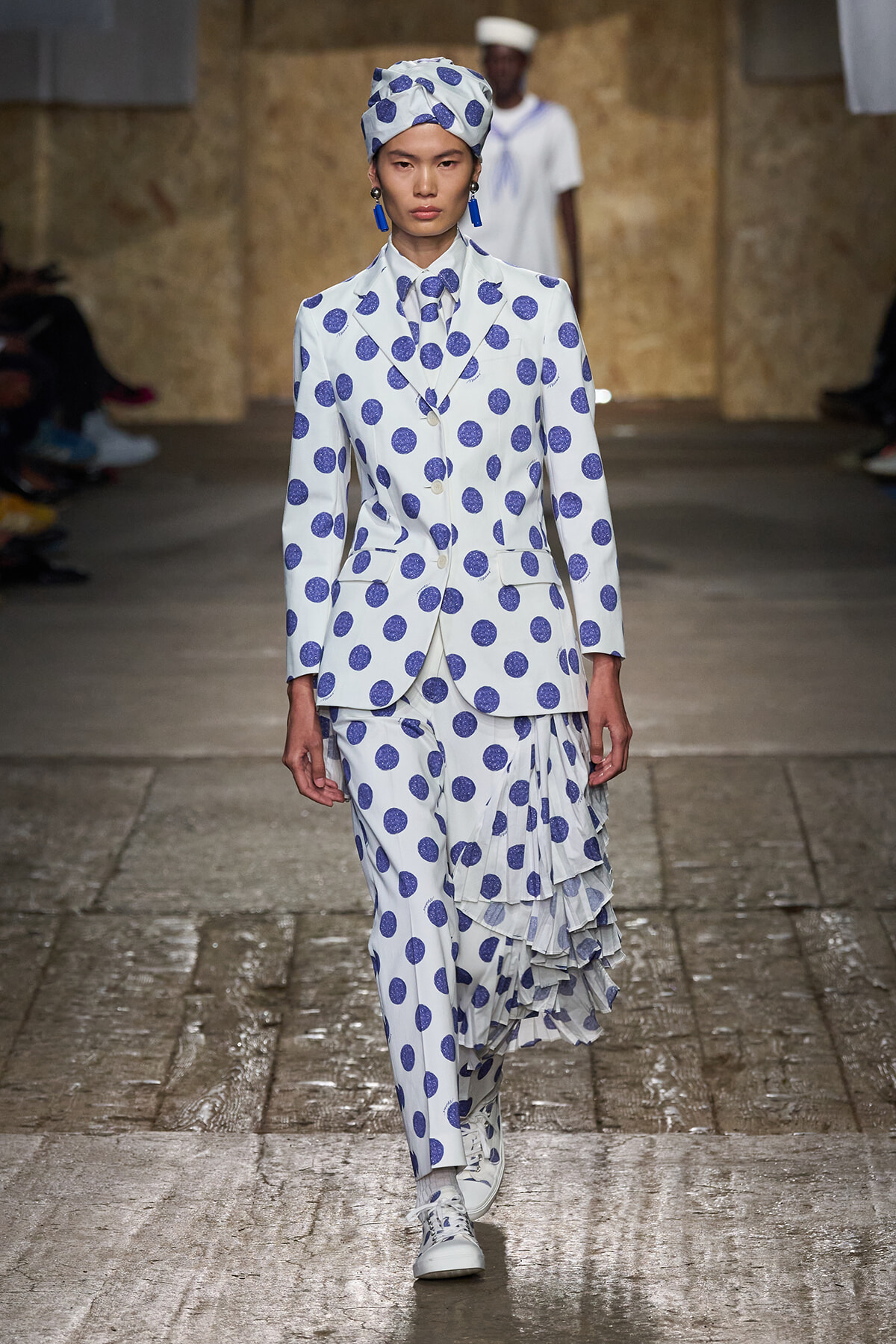 Model walks a runway in a white suit with blue polka dots, matching headscarf, and a layered_ruffled skirt, white sneakers visible preppy look.