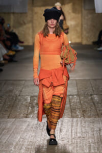 Model on a runway wearing an orange-toned, layered outfit with a black knit beanie and a fringed tan bag.
