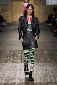 Model walks a fashion runway in a black leather biker jacket and matching skirt, with graphic green-printed leggings and black lace-up boots.