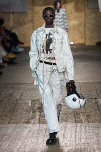 Runway model in a light-washed denim jacket and jeans with a graphic tee, oversized pearl necklaces, round sunglasses, and black boots, carrying a white helmet.