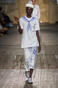 Model on a runway wearing a white tunic with a blue sailor knot graphic and matching white pants with blue illustrations, plus a white hat and blue earrings.