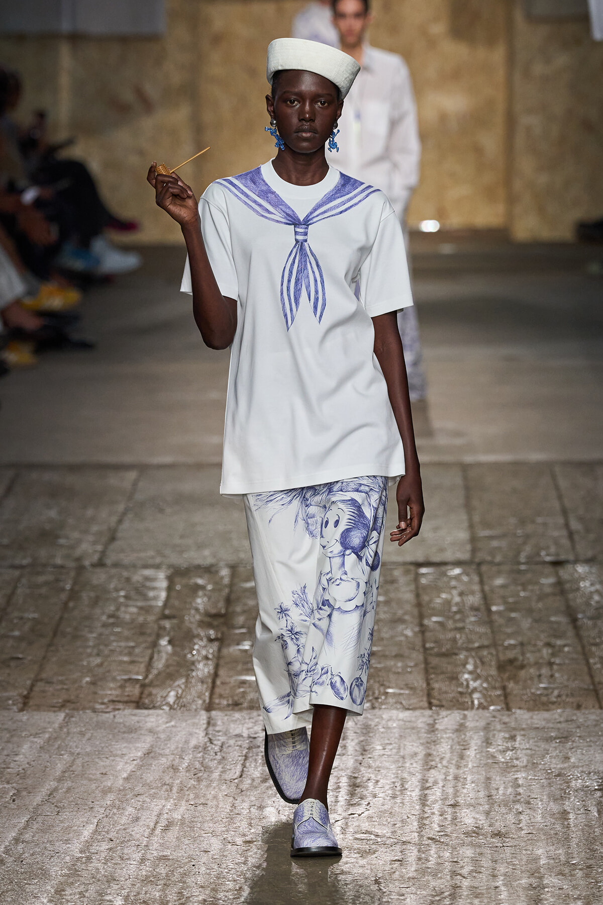 Model on a runway wearing a white tunic with a blue sailor knot graphic and matching white pants with blue illustrations, plus a white hat and blue earrings.