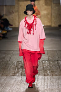 Runway model in a pale pink top, vivid red pants, a chunky red necklace, and a black bucket hat.