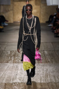 Runway model in a black dress draped with pearl strands, with pink and yellow lace at the hem, black boots, and a bright pink handbag.