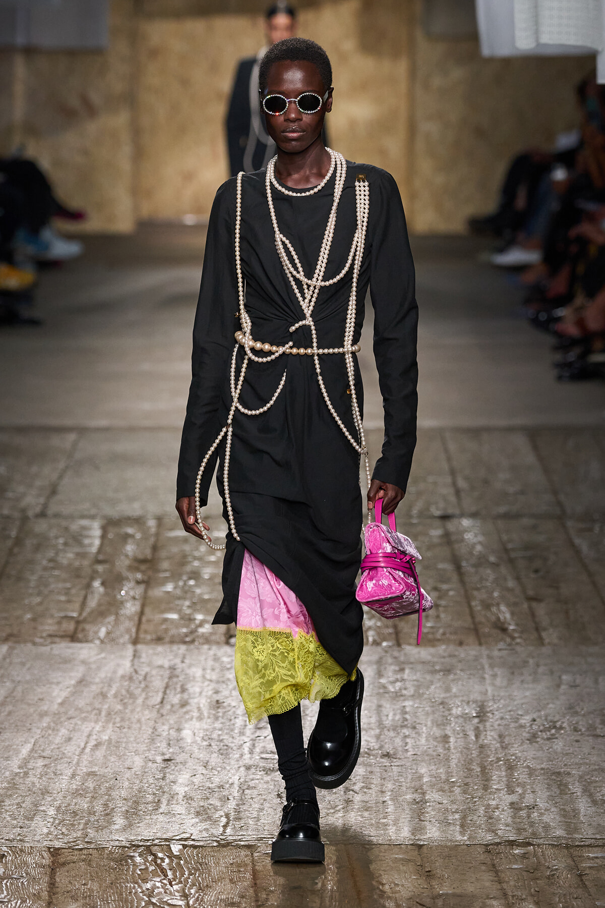 Runway model in a black dress draped with pearl strands, with pink and yellow lace at the hem, black boots, and a bright pink handbag.