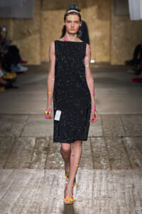 Model walks the runway in a knee-length black sleeveless dress sprinkled with small reflective threads and multicolored confetti stickers on arms, legs, and feet.