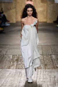 Model walks the runway in a white strapless, draped dress with a front knot and layered skirt, hands in pockets.