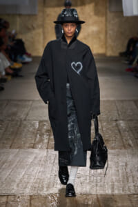 Model walks the runway in a black oversized coat with a white heart chalk sketch, paired with a black wide-brim hat and bag.