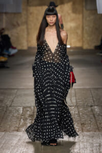 Model walks the runway in a sheer black polka-dot gown with a deep V neckline, carrying a pearl-strap bag.