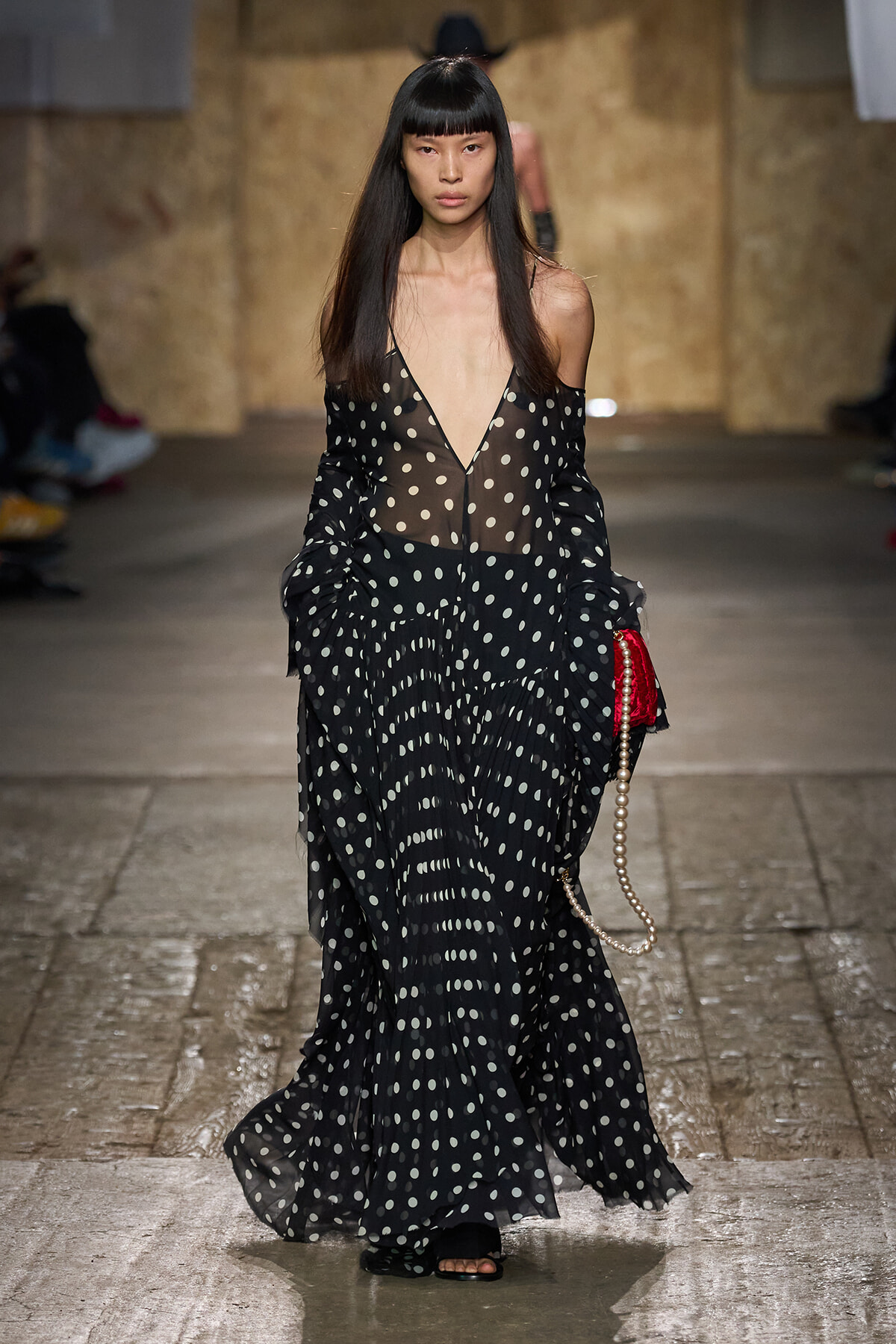 Model walks the runway in a sheer black polka-dot gown with a deep V neckline, carrying a pearl-strap bag.
