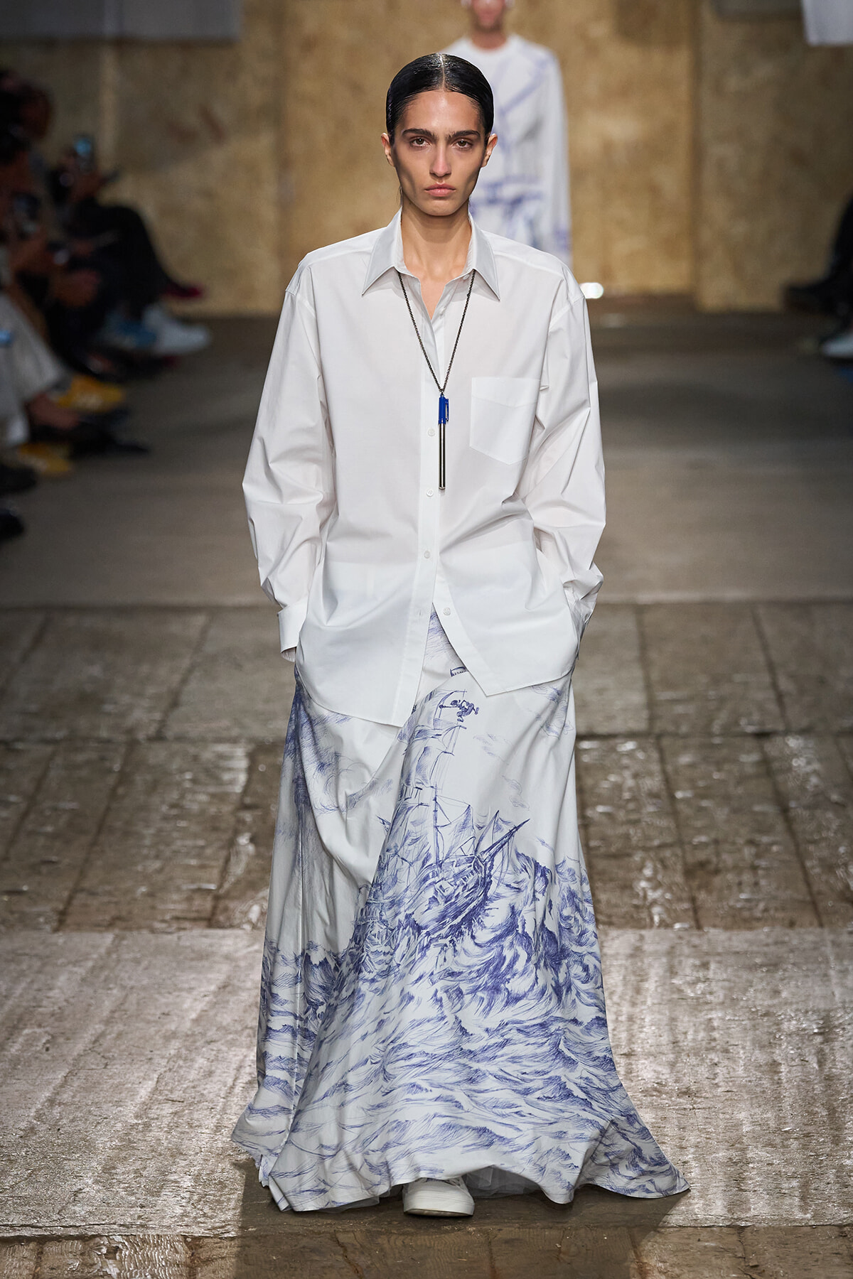 Model walks the runway in a loose white button-down shirt paired with a long white skirt featuring blue nautical-wave print and a pendant necklace with blue accents.
