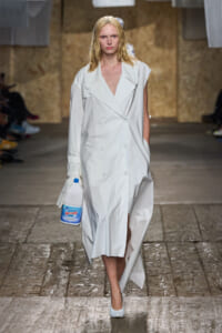 Model walking runway in a long oversized white trench coat, holding a large blue-and-white water jug in her gloved hand.