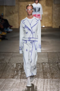Female model walks a runway in a white trench coat with blue sketch-print lines, white sneakers, and bold white glasses.