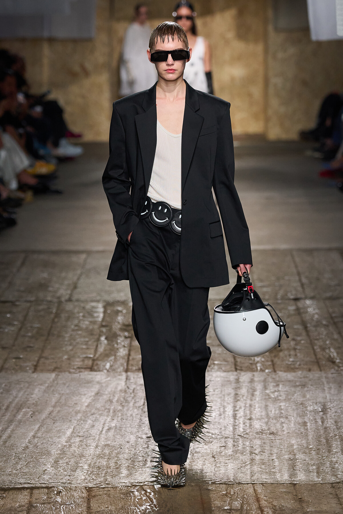Model on a runway wearing a black suit over a white top, oversized blazer, spiked heels, and a white helmet with black detailing.