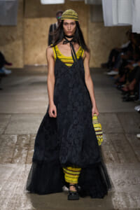Model on a runway wearing a yellow striped top under a black floral dress with a sheer train, a yellow/black striped beanie, and a yellow striped clutch.