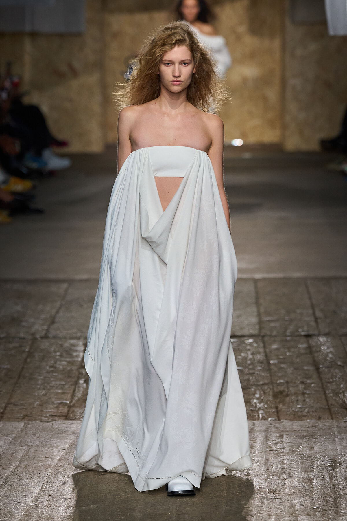 Model walking a fashion runway in a white draped gown with a strapless bodice and cutout at the midsection.