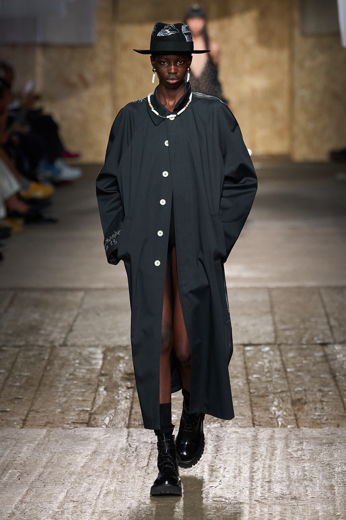 Model walking a fashion show in a long dark coat, black hat, and chunky boots on a wet runway.