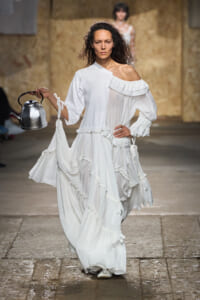 Model walking the runway in a flowing white off-shoulder dress with ruffles, holding a metal kettle in her left hand.