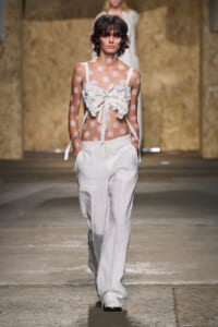Runway model in a sheer polka-dot top with oversized white ruffles and white wide-leg pants.
