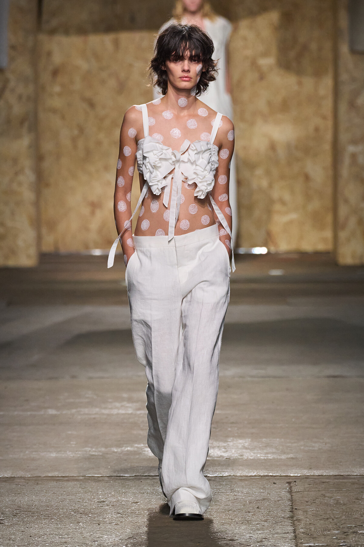 Runway model in a sheer polka-dot top with oversized white ruffles and white wide-leg pants.