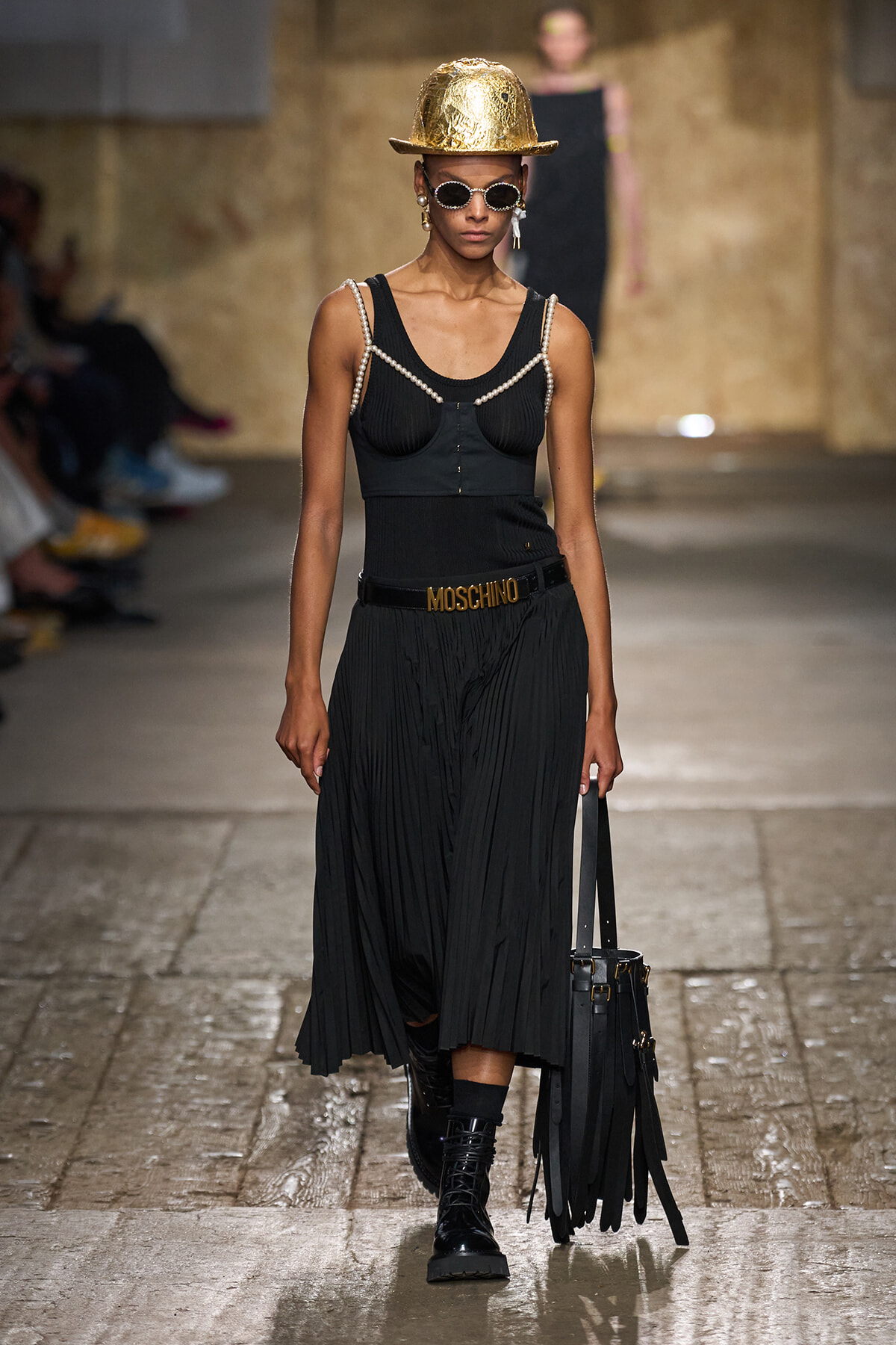 Female model on a runway wearing a black pleated skirt and corset-style top with pearl straps, a gold helmet, sunglasses, and a fringed black handbag with a Moschino belt.