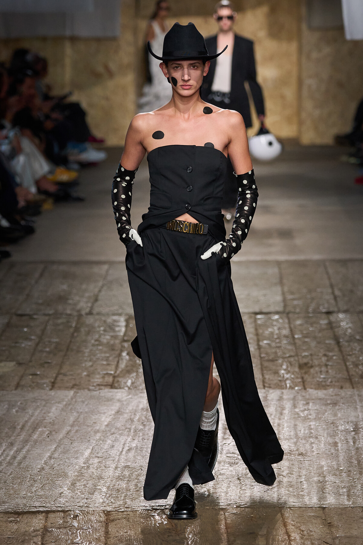 Model walks the runway in a black strapless top and wide-leg skirt, wearing a black cowboy hat, polka-dotted long gloves, and a Moschino belt with bold dot makeup accents on the face and shoulders.